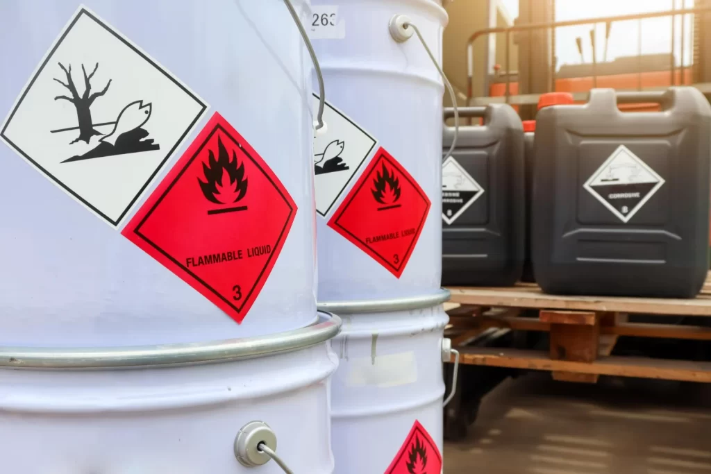 Dois tanques químicos brancos com um símbolo de líquido inflamável, representando a indústria de transporte de cargas perigosas. A imagem destaca a sinalização de segurança, essencial para a identificação de produtos químicos e substâncias perigosas durante o transporte. Ao lado, outros galões pretos com diferentes tipos de carga perigosa estão sobrepostos a um deck de madeira.