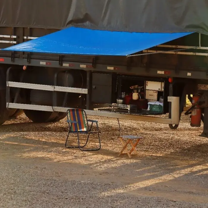 Toldo de lona azul instalado na lateral do caminhão criando área de sombra com cadeira e mesa